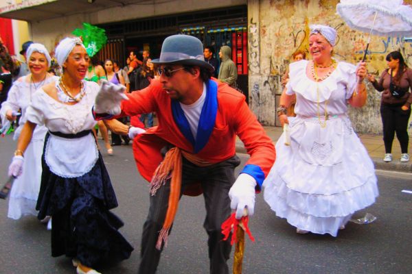 Carnival scene from the streets of San Telmo, Buenos Aires, with man in bowler hat and women in large dresses and umbrellas.