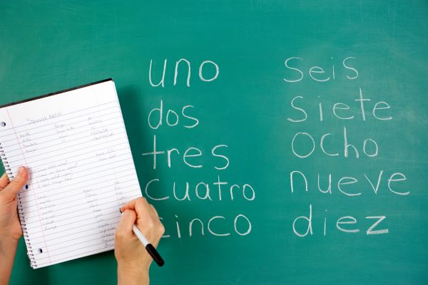 Image of hand taking notes on a notepad ontop of a chalkboard with numbers written in Spanish.