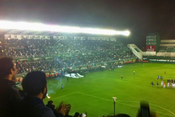 Nighttime football match at CA Banfield stadium in Buenos Aires, Argentina, with players on the field under bright floodlights and a crowd of passionate fans in the stands.