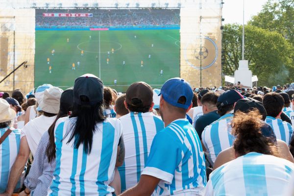 Fans of Argentina gathered outdoors, facing a large screen, cheering for their football team."